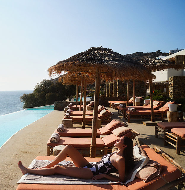 A woman sunbathing on a lounger with a luxury Sun of a Beach towel at a premium poolside, featuring stunning views of the sea and mountains.
