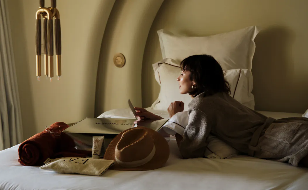 A woman is comfortably nestled in a luxurious bathrobe, engrossed in reading the welcome note that has been thoughtfully placed for her. Surrounding her on the bed are a stylish hat, a soft towel, a soothing cream, and a chic pouch, creating a serene atmosphere.
